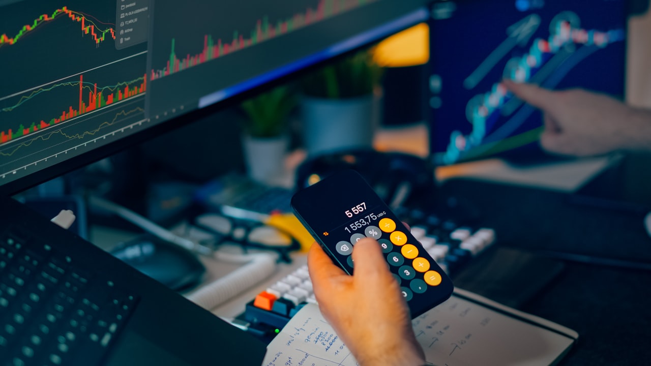 Trader analyzing stock market charts on computer screens with calculator.